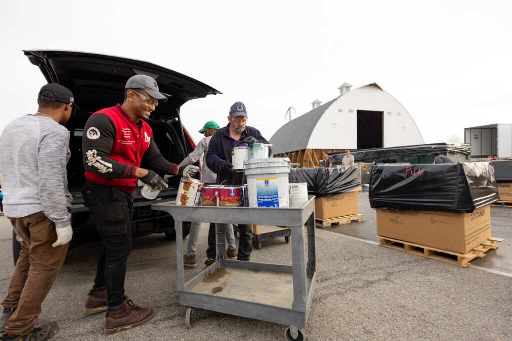 Four workers unloading paint cans from a car trunk onto a cart outside a white barn-like building.