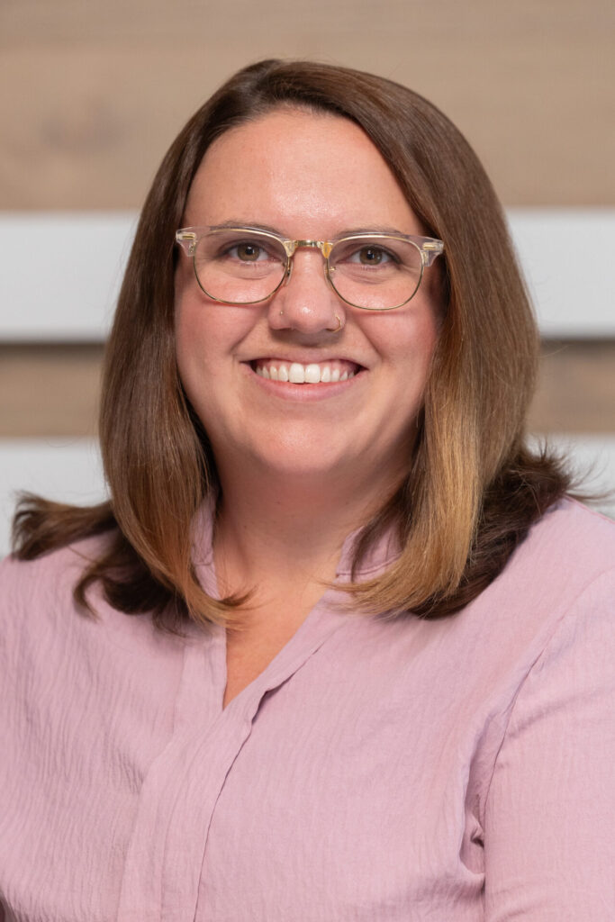 Smiling woman with shoulder-length brown hair, clear-framed glasses and a nose ring, wearing a light pink blouse.