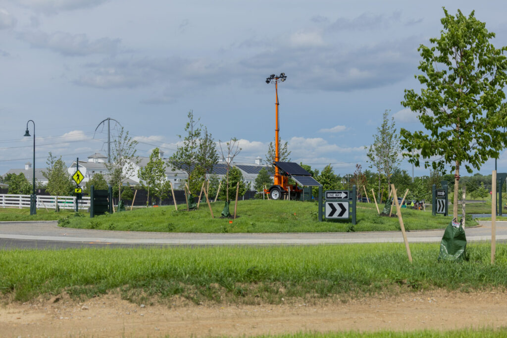 Roundabout with grassy center, young trees, an orange trailer-mounted light tower with solar panels, and directional signs