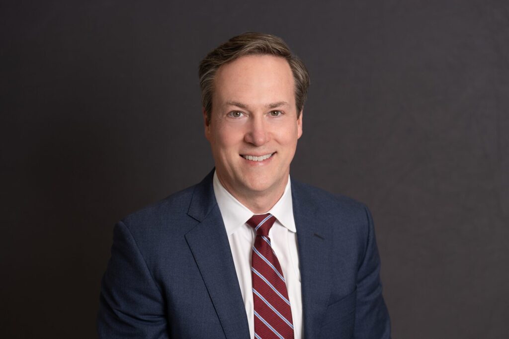 Smiling middle-aged man in a navy suit, white shirt and red striped tie against a dark gray studio background.