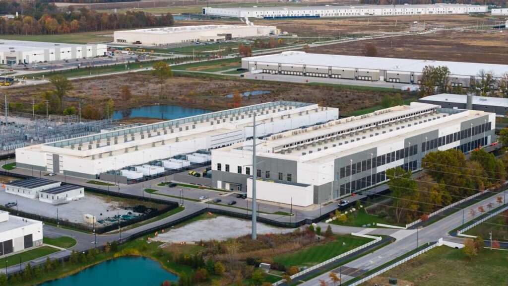 Large modern data center or warehouse campus with long white buildings, rooftop cooling units, parking areas and surrounding service roads and ponds