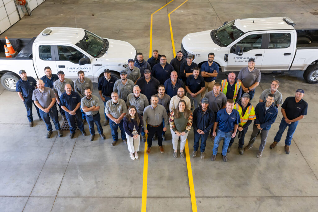 Group of city public works employees standing in a garage between two white New Albany Public Service trucks, smiling up at camera