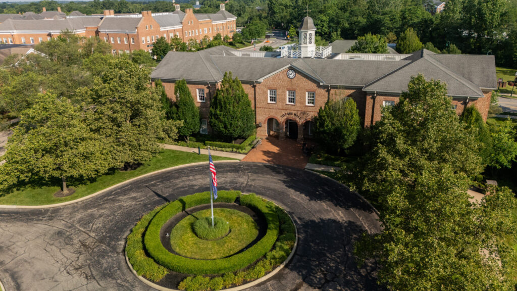 Brick village hall with clock tower behind circular driveway and flagpole, surrounded by trees and manicured hedges
