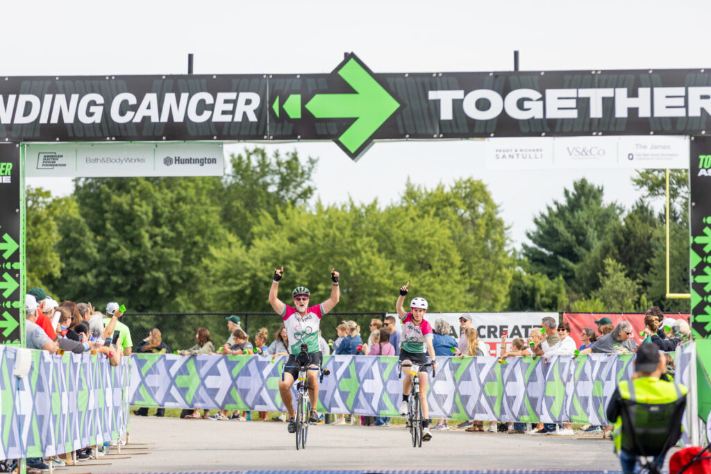 Two bike riders cross the finish line with their hands up in celebration.