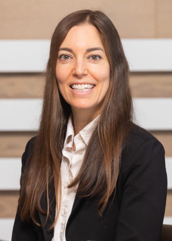 Alexandra Fox smiles for a headshot. She is wearing a black jacket and white shirt and is sitting in front of a brown backdrop.