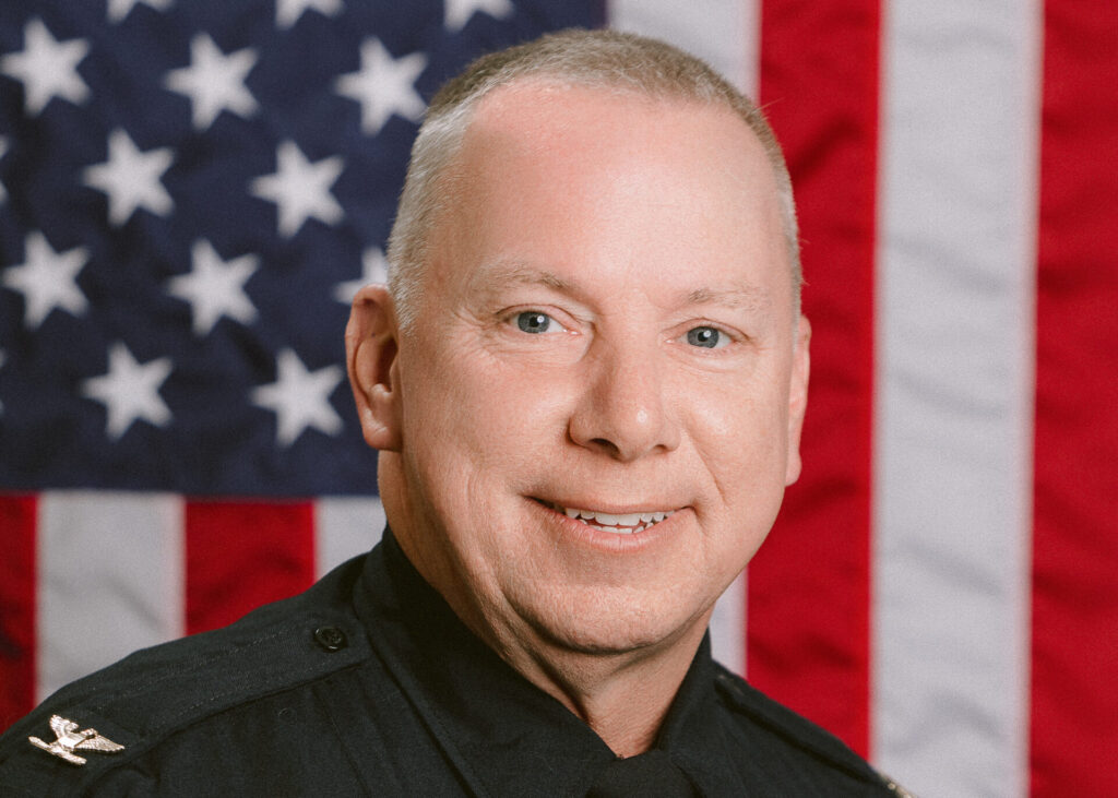 Greg Jones smiles for a headshot. He is wearing his police uniform and is in front of an American Flag