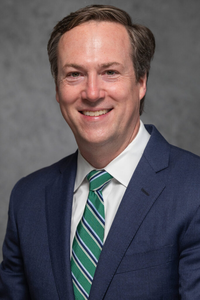 John McClelland Smiles for a headshot. Wearing a Navy Blue suit and green tie with a dark gray background.