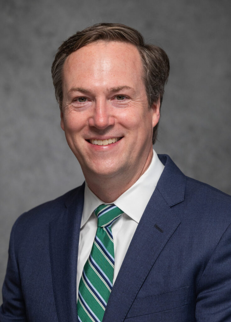 John McClelland Smiles for a headshot. Wearing a Navy Blue suit and green tie with a dark gray background.