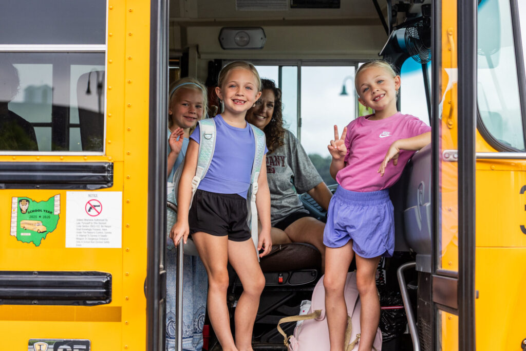 Three students wearing backpacks stand in the steps of a school bus with the school bus driver.