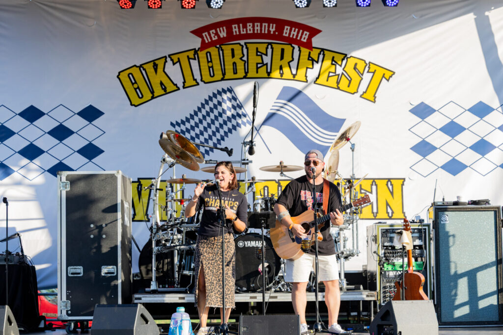 Two musicians standing on a stage performing. The backdrop reads "New Albany, Ohio Oktoberfest"