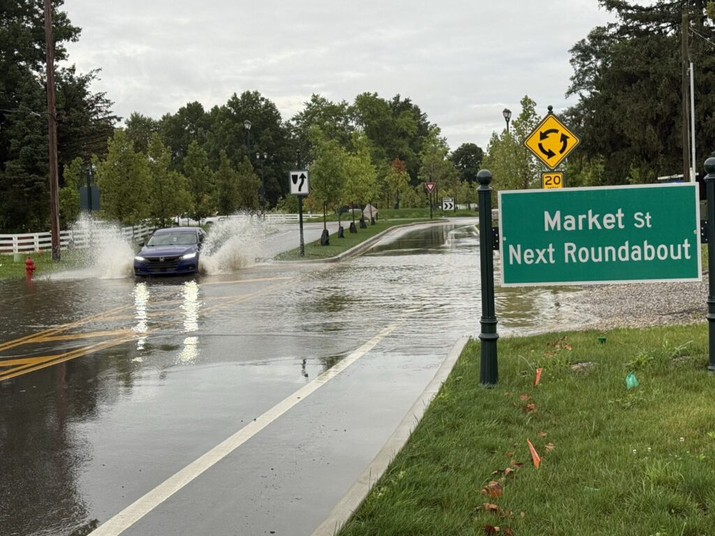 A blue car driving through floods with a green sign saying "Market St. Next Roundabout"