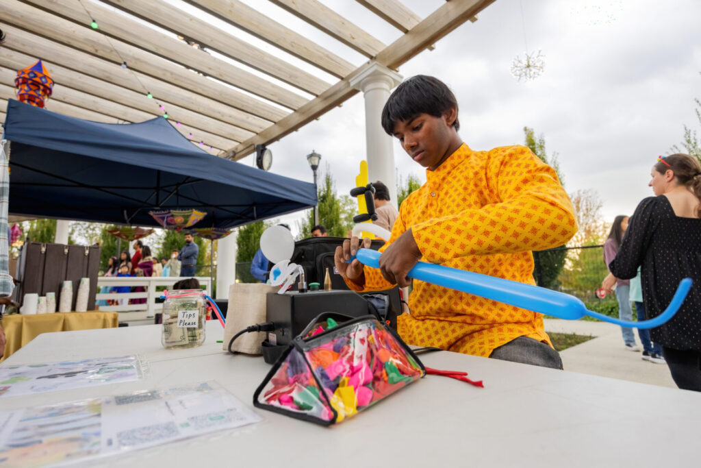 A vendor is creating balloon art at a table. the sky is gray and the vendor is wearing traditional wear