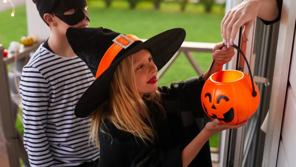 Two people in Halloween costumes at a doorway, one in a striped shirt and black mask, the other in a black-and-orange witch hat holding an orange jack-o’-lantern bucket with a handle.