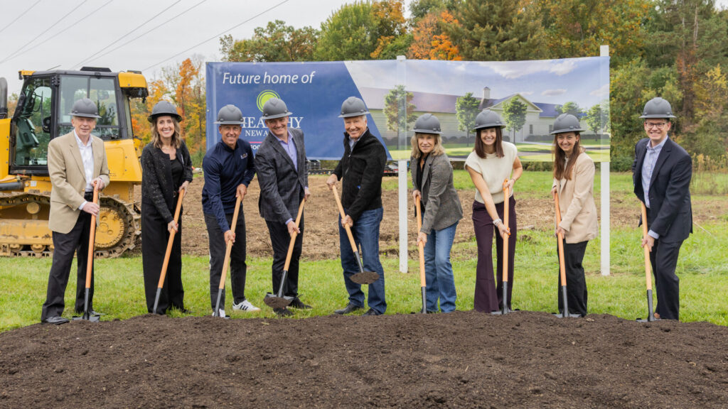 9 people with hard hats and shovels dig dirt at a ground breaking.